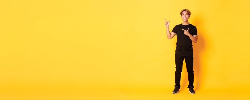 Full-length of smiling attractive korean guy pointing fingers left, showing logo, yellow background.