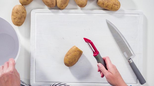 Flat lay. Step by step. Peeling large yellow potatoes with a vegetable peeler on a white wooden board.