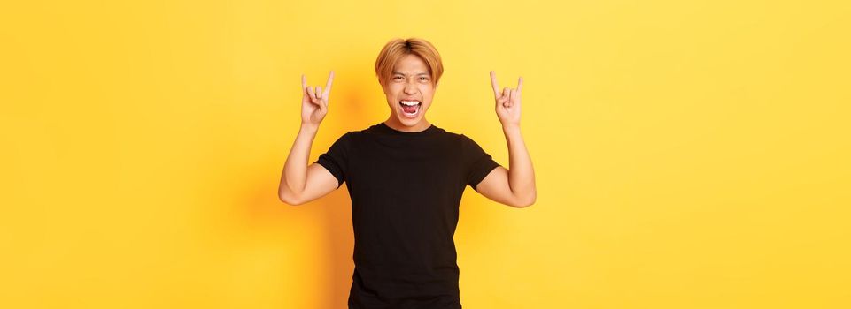 Portrait of happy handsome asian man having fun and partying, showing rock-n-roll gesture, standing yellow background.