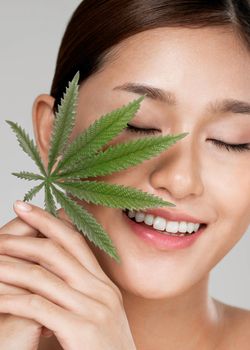 Closeup portrait of young ardent girl with healthy fresh skin holding green hemp leaf. Combination of beauty and cannabis concept.