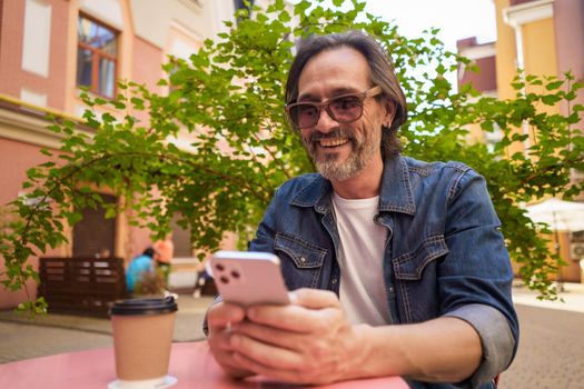 Middle aged handsome freelancer man texting or having a work video call while enjoying coffee outdoors. Mature man looking at phone in hands sitting at cafe while working outdoors.