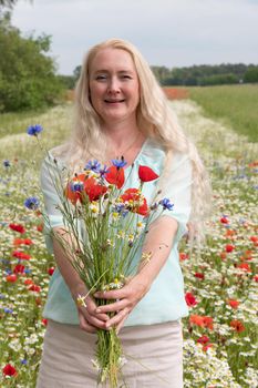 a beautiful middle-aged blonde woman stands among a flowering field of poppy, daisies, Cornflowers, and holds a bouquet of wild flowers and laughs. High quality photo