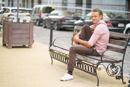 Young handsome man sits on the bench with one leg up on it spend time in urban city near his office or home, wearing pink shirt and shoulder bag. Freelancer man off work.