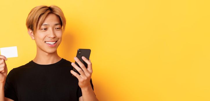 Close-up of handsome stylish asian guy shopping online, looking at mobile phone and smiling, showing credit card, standing over yellow background.