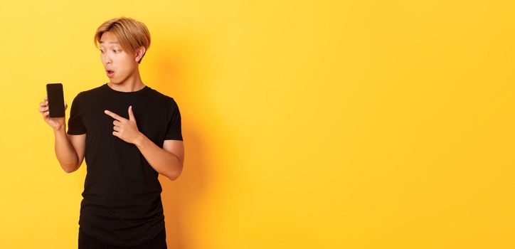 Portrait of handsome asian man showing something on smartphone screen, pointing at mobile phone display, standing over yellow background.