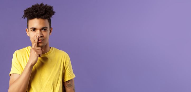 Close-up portrait of funny and unappropriate young hispanic teenage guy picking nose with finger, smiling and looking at camera, standing purple background, have bad habit.