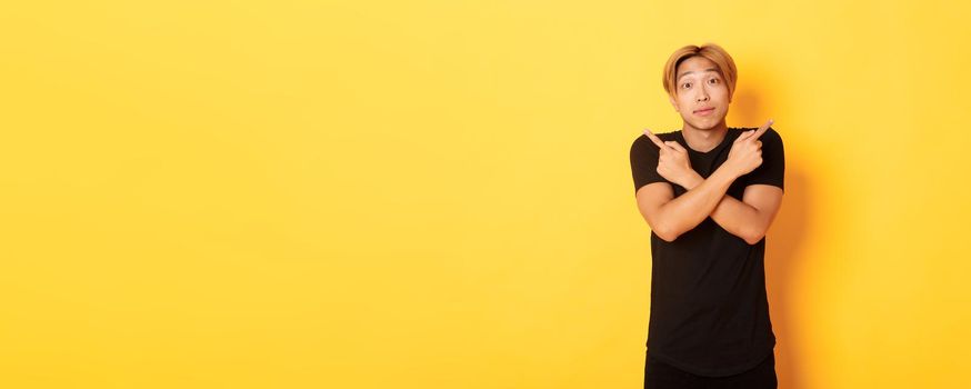 Indecisive handsome guy shrugging puzzled and pointing fingers sideways, standing yellow background.