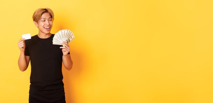 Happy and lucky asian guy looking pleased at money while showing credit card and cash, standing yellow background.