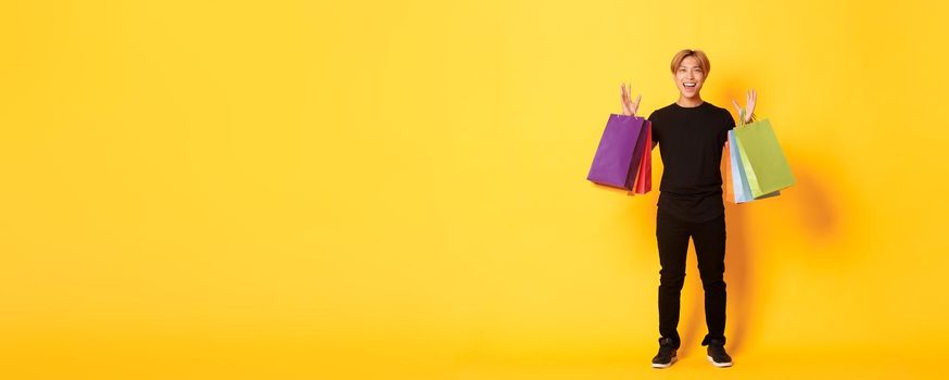 Full-length of happy handsome asian guy on shopping, holding bags and smiling, yellow background.