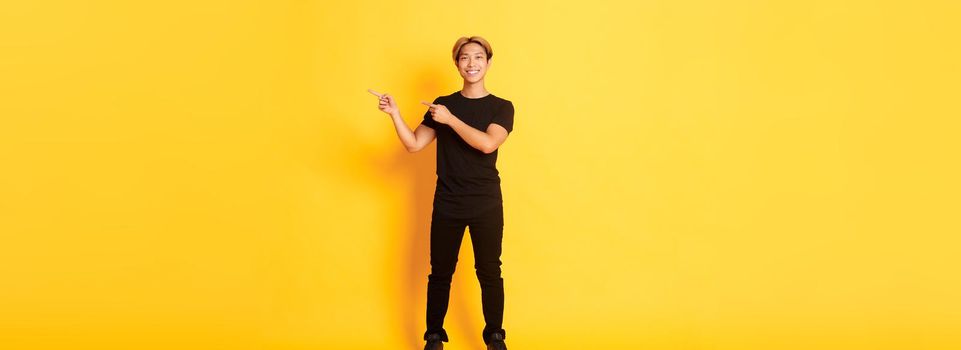 Full-length of handsome confident asian guy smiling happy, pointing fingers right, showing logo, yellow background.