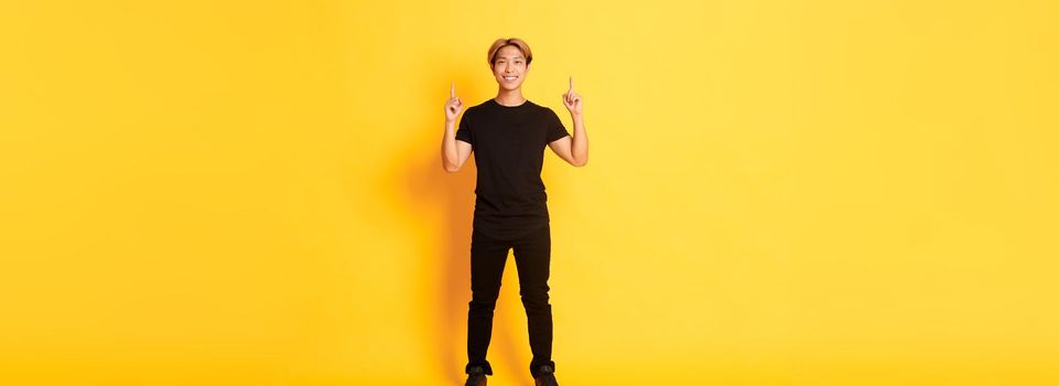 Full-length of confident handsome asian man with blond hair, smiling and pointing fingers up, showing logo, yellow background.
