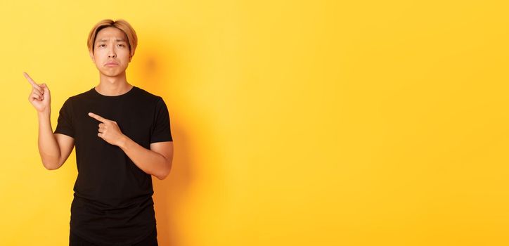 Portrait of misearble and gloomy asian guy in black t-shirt, pointing fingers upper left corner disappointed, yellow background.