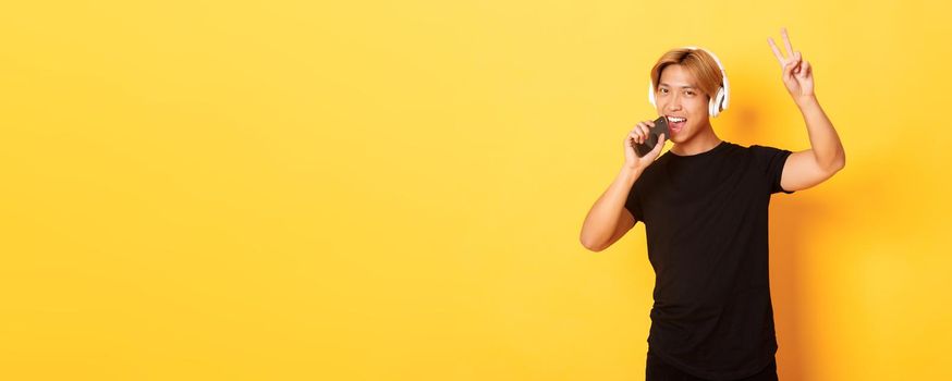 Sassy handsome asian guy in headphones, playing karaoke app, singing into mobile phone microphone, showing peace gesture, standing yellow background.