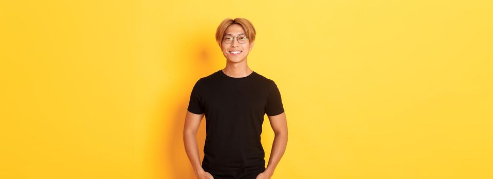 Handsome asian blond guy in glasses standing in black clothes and smiling pleased, yellow background.