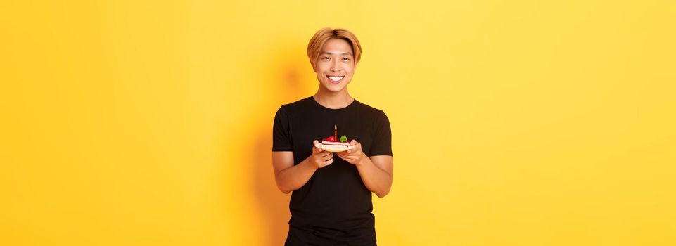 Handsome happy asian blond guy, smiling pleased as celebrating birthday, holding b-day cake, standing over yellow background.