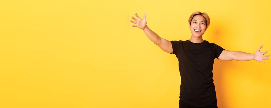 Portrait of handsome asian guy spread hands for hug, cuddling someone friendly, standing yellow background.