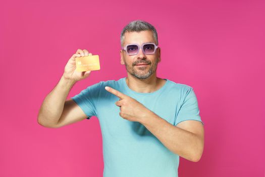 Serious handsome middle aged man hold debit, credit card in hand pointing at it wearing blue t-shirt and glasses isolated on pink background. Financial, banking concept.