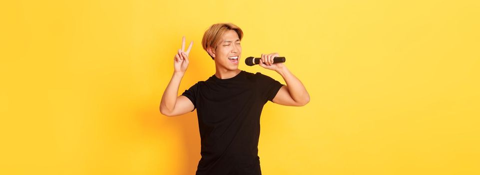 Carefree handsome asian guy performing song, singing into microphone and showing peace gesture, standing yellow background.