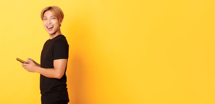 Portrait of handsome stylish asian guy in black outfit, using mobile phone and turning head at camera with satisfied smile, yellow background.