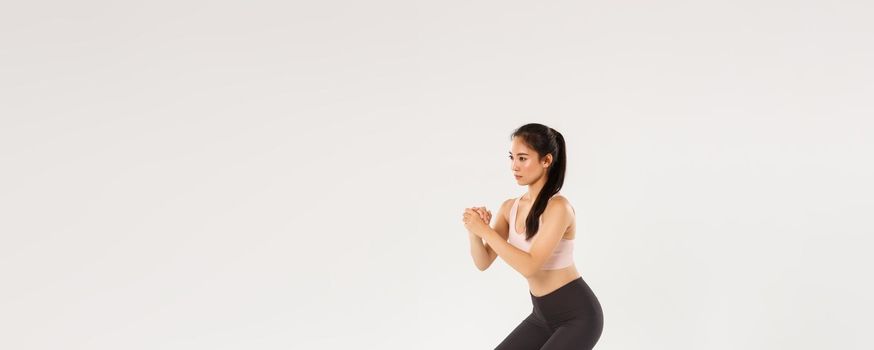 Full length side view of focused slim asian girl doing fitness training, female athlete clasp hands together and perform squats exercises with stretching resistance band, workout equipment.