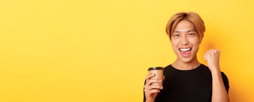 Close-up of energized handsome asian guy fist pump joyfully while drinking coffee, smiling excited over yellow background.