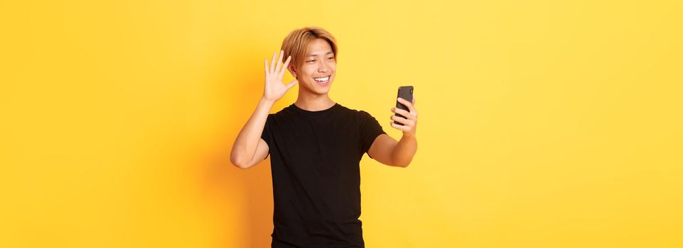 Portrait of friendly handsome asian man, smiling and waving hand at smartphone camera, greeting friends on video call, standing yellow background.