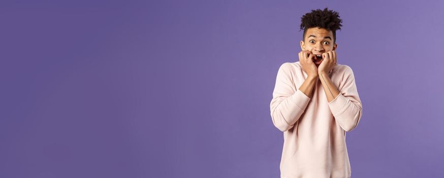 Portrait of young scared guy watching really scary movie, horror alone, gasping screaming and hold hands pressed to face, look terrified, trembling from fear, standing purple background.