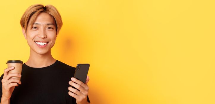 Close-up of handsome young asian guy smiling happy, using smartphone and drinking takeaway coffee, standing over yellow background.