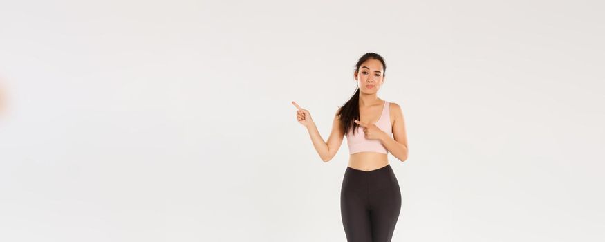 Full length of skeptical and doubtful asian female brunette, girl athlete have doubts, pointing fingers upper right corner, frowning and looking with disbelief, sportswoman showing promo.