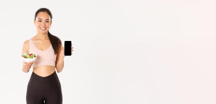 Sport, wellbeing and active lifestyle concept. Smiling slim and cute asian fitness girl, gym coach showing salad and smartphone screen, recommend download diet tracker or calories reminder.