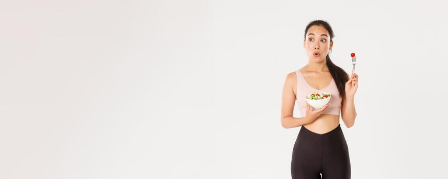 Sport, wellbeing and active lifestyle concept. Amused and wondered cute slim asian fitness girl in activewear looking left at your logo or advertisement while eating salad, white background.