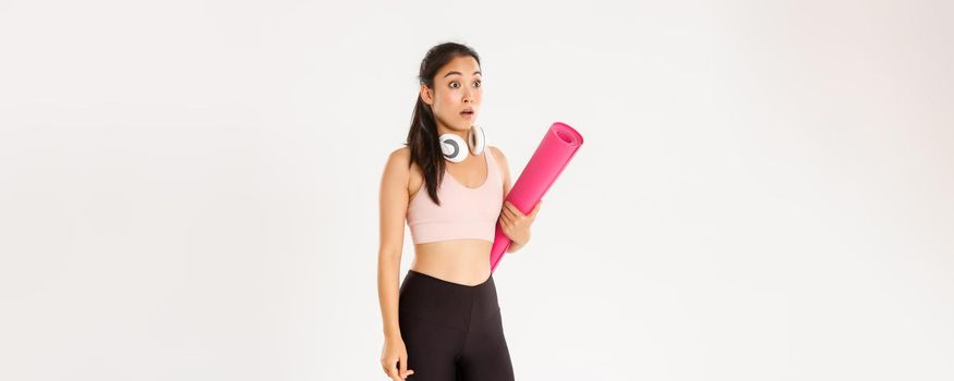 Sport, wellbeing and active lifestyle concept. Shocked asian fitness girl in stupor, drop jaw and gasping while looking left at logo or banner info, holding rubber mat for fitness exercises.