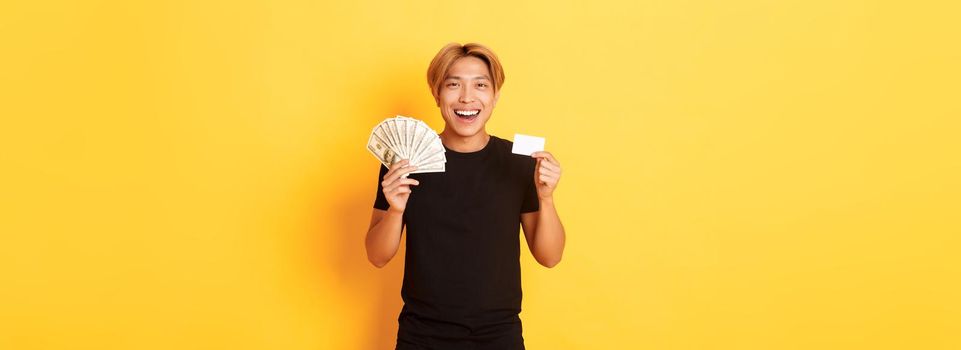 Young happy asian man with blond hair, showing credit card and money, smiling cheerful, standing yellow background.