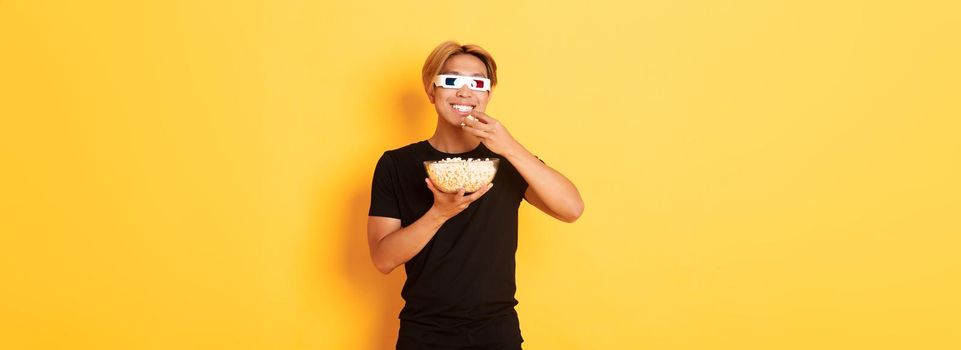 Joyful handsome asian guy with blond hair, watching movie or tv series in 3d glasses, eating popcorn and smiling amused, standing yellow background.