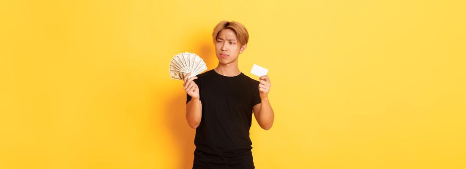 Indecisive and confused young asian guy looking puzzled at money while holding cash and credit card, yellow background.