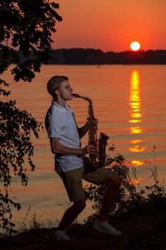 A young guy plays the saxophone in the evening at sunset.