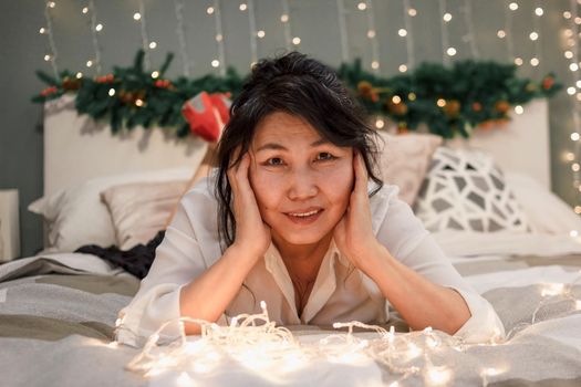 Senior asian woman relax on bed with xmas lights looking at camera, portrait of charming elderly smiling lady in bedroom at home at christmas holiday