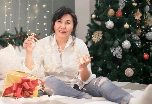 Senior happy asian woman on bed with xmas lights looking at camera, portrait of attractive elderly smiling lady with garland in hands and gold gift at home at christmas holiday, atmospheric mood