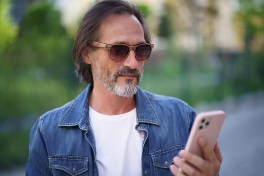 Middle aged man looking at smartphone standing on street while enjoying free time off work. Mature man read travel digest using mobile phone traveling old town. Business on the go.