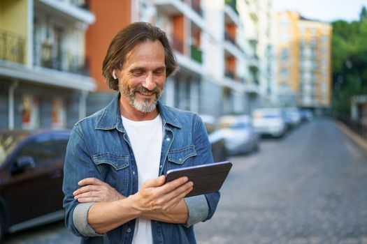 Traveling handsome mature man looking at digital tablet in hand talking outdoors having a call on urban city streets. Mature man listening music use wireless earphones while travel old town streets.