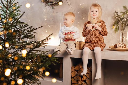 Little brother and sister play on Christmas eve in a beautiful house decorated for the New Year holidays. Children are playing with a Christmas gift. Scandinavian-style interior with live fir trees and a wooden staircase.