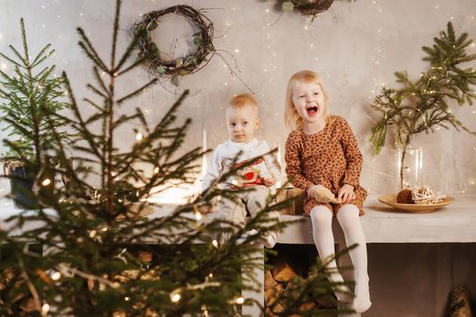 Little brother and sister play on Christmas eve in a beautiful house decorated for the New Year holidays. Children are playing with a Christmas gift. Scandinavian-style interior with live fir trees and a wooden staircase.