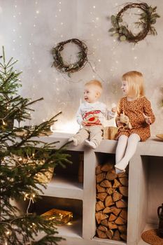 Little brother and sister play on Christmas eve in a beautiful house decorated for the New Year holidays. Children are playing with a Christmas gift. Scandinavian-style interior with live fir trees and a wooden staircase.