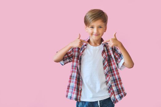Young happy boy points to his clothes with thumb up, isolated on pink background with copy space. Blank white t-shirt for your design