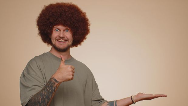 Handsome man in t-shirt showing thumbs up and pointing empty place, advertising area for commercial text, copy space for goods promotion. Young adult guy. Indoors studio shot on beige wall background