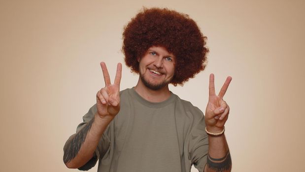 Bearded man with lush hairstyle showing victory sign, hoping for success and win, doing peace gesture, smiling with kind optimistic expression. Young adult guy boy isolated on beige studio background