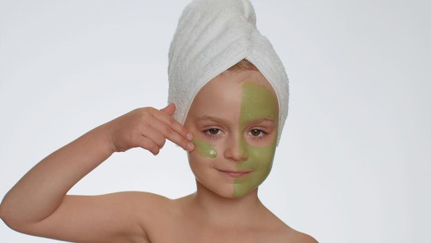 Beautiful young smiling child girl in towel on head applying cleansing moisturizing green facial mask. Teenager kid face skin care treatment, natural cosmetics. Female portrait. Perfect fresh clean