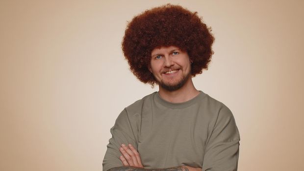 Cheerful lovely bearded adult man in casual t-shirt smiling, looking at camera. Young fashion model guy boy with lush hairstyle coiffure indoor studio isolated on beige background. Male nature beauty