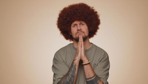 Please, God help. Sad worried man praying, looking upward, making wish asking with hopeful imploring expression begging apology, forgiveness. Young adult guy indoor isolated alone on beige background