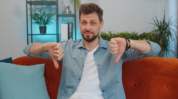 Dislike. Upset man showing thumbs down sign gesture, expressing discontent, disapproval, dissatisfied bad work at modern home apartment indoors. Displeased young guy in living room sitting on sofa